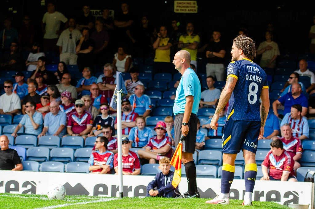 Southend player stands beside the assistant referee at the corner flag with Scunthorpe supporters behind them.